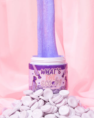 A jar of 'What's the Scoop?' stretchy body scrub by P.Louise is displayed with an array of parma lilac sweets in the foreground and the body scrub being stretched out of the pot, against a pink backdrop.